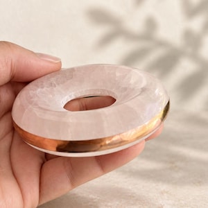 May include: A pale pink rose quartz stone shaped like a smooth donut or ring. The stone features a translucent, crystalline texture and is accented with a polished metallic gold band encircling the outer edge of the circular form.