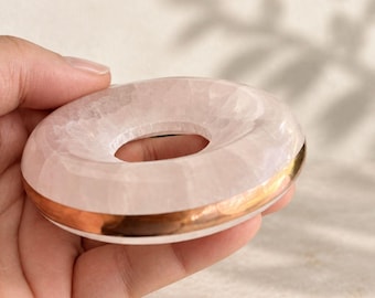 May include: A pale pink rose quartz stone shaped like a smooth donut or ring. The stone features a translucent, crystalline texture and is accented with a polished metallic gold band encircling the outer edge of the circular form.