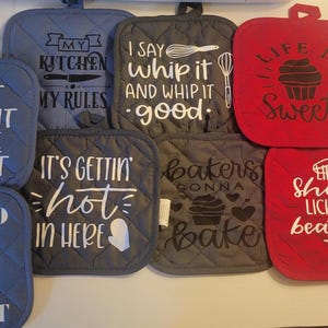 Op de afbeelding: Set van tien grijze en rode gewatteerde ovenwanten met verschillende spreuken met een keukenthema. De spreuken zijn: "Laat het niet vallen, want het is heet", "Mijn keuken, mijn regels", "Ik zeg kloppen en kloppen goed", "Het leven is zoet", "Het wordt hier heet", "Bakkers gaan bakken", "Het leven is kort, lik de kloppers", en "Hier om de pot te roeren".