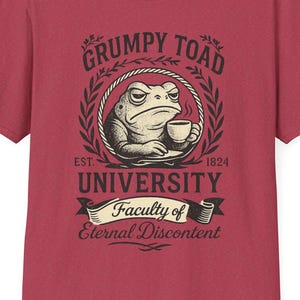 May include: A maroon t-shirt featuring a graphic of a grumpy toad holding a cup of coffee, encircled by a laurel wreath. The text reads "Grumpy Toad University, Faculty of Eternal Discontent."