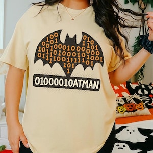 May include: A light yellow t-shirt featuring a black bat silhouette filled with binary code. Below the bat, the text "01000010ATMAN" is displayed in a black rectangle. The shirt is paired with black shorts, suggesting a casual outfit.
