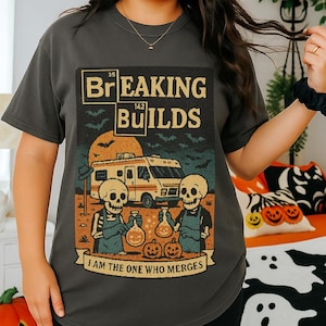 May include: Dark gray t-shirt with the text "Breaking Builds" and a graphic of skeletons, a camper van, and pumpkins. The shirt has a vintage, Halloween-themed design with the phrase "I am the one who merges".