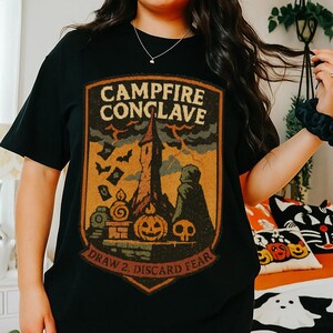 May include: Black t-shirt with a Halloween-themed graphic. The design features a spooky castle, bats, pumpkins, and the text "Campfire Conclave" and "Draw 2, Discard Fear". The shirt is paired with black shorts, perfect for a casual fall outfit.