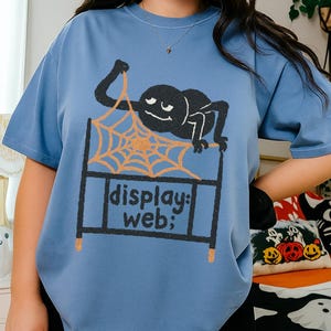 May include: A light blue t-shirt featuring a black spider hanging from an orange web. The web is above a black rectangle with the words "display: web;" in white. The shirt is a casual, oversized fit.