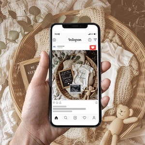 May include: A smartphone displaying an Instagram post announcing a pregnancy with the text "We have been keeping a little secret... Arriving 2023" and a photo of a baby onesie with the text "Baby Coleman" and an ultrasound image. The post is set against a background of a wicker basket filled with eucalyptus leaves and a wooden toy bear.