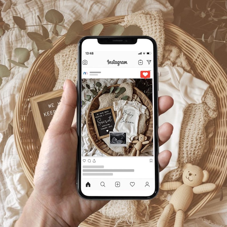 May include: A smartphone displaying an Instagram post announcing a pregnancy with the text "We have been keeping a little secret... Arriving 2023" and a photo of a baby onesie with the text "Baby Coleman" and an ultrasound image. The post is set against a background of a wicker basket filled with eucalyptus leaves and a wooden toy bear.