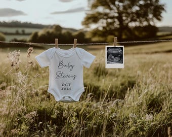May include: A white baby onesie with the text "Baby Stevens OCT 2025" printed on it. A black and white ultrasound image is hanging next to it.