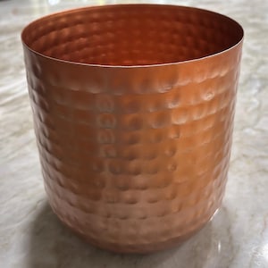 May include: A copper-colored, cylindrical container with a hammered texture. The vessel has a wide opening and a rounded base. The copper finish reflects light, creating highlights and shadows across the surface. The container is likely used for storage or decoration.