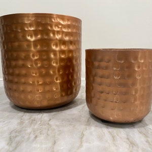 May include: Two hammered copper planters, one larger than the other, with a textured surface. The planters are a warm copper color.