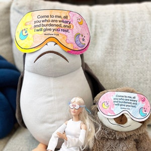 May include: Two plush toys wearing eye masks with the text "Come to me, all you who are weary and burdened, and I will give you rest. Matthew 11:28" printed on them. A doll with blonde hair is sitting in front of the toys.
