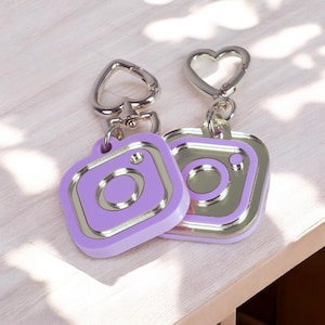 Op de afbeelding: Twee vierkante sleutelhangers met een gestileerd camerapictogram in lavendel en metallic zilver. Elke sleutelhanger heeft een hartvormige metalen sluiting aan de bovenkant. Het ontwerp bootst het logo van een social media camera na.