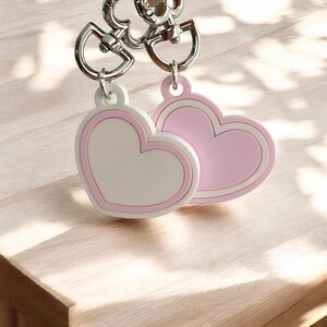 May include: Two heart-shaped charms attached to silver-toned metal swivel clasps. One charm features a white center with a light pink border, while the other charm is solid light pink with a matching pink outline. Both charms have a smooth finish.