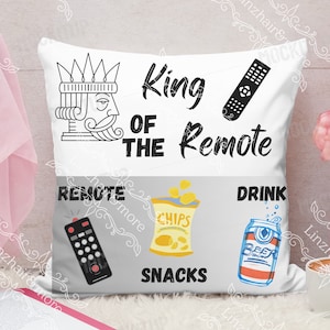 May include: A white throw pillow with black text that reads "King of the Remote". The pillow has a graphic of a king's head with a crown, a remote control, a can of beer, a bag of chips, and a remote control. The pillow is sitting on a white surface with a book and a cup of coffee.