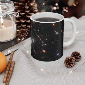May include: White ceramic mug with a black and white image of a galaxy with stars and nebulae. The mug is filled with coffee and steam is rising from the top.