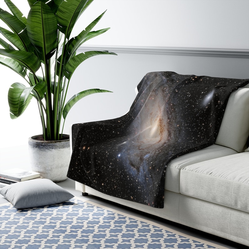 Pu&ograve; includere: Una coperta nera e bianca con una stampa di galassia che presenta una nebulosa vorticosa. La coperta &egrave; drappeggiata su un divano bianco con una pianta verde in un vaso in primo piano.