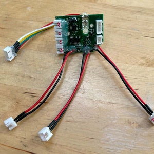 Könnte beinhalten: Eine grüne Leiterplatte mit verschiedenen elektronischen Bauteilen, Drähten und Steckern. Die Platine hat mehrere weiße Stecker und Drähte in Rot, Schwarz, Gelb und Weiß. Die Komponenten sind auf einer Holzoberfläche angeordnet.