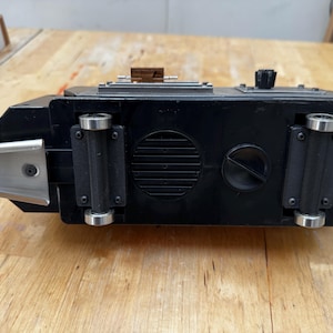 May include: A black rectangular device with metal accents, a knob, and small wheels. The device has a silver metal bracket on one end and a series of vents and a circular cover on the bottom. The device is resting on a wooden surface.