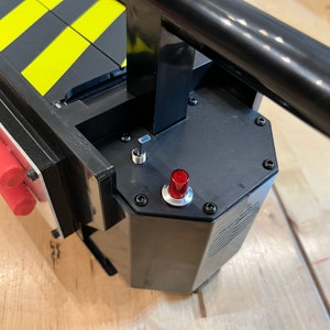 May include: A black and yellow Ghostbusters proton pack with a red button and a black lever. The pack has a black handle and a black and yellow striped design.