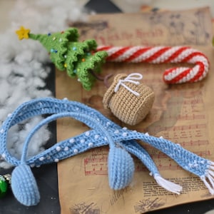 May include: A blue crocheted headband with white polka dots and a matching blue crocheted ear warmer. The headband is lying on a piece of paper with music notes and a crocheted candy cane, Christmas tree, and small bag.