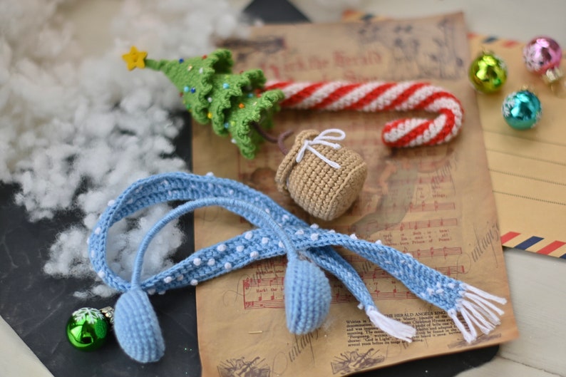 May include: A blue crocheted headband with white polka dots and a matching blue crocheted ear warmer. The headband is lying on a piece of paper with music notes and a crocheted candy cane, Christmas tree, and small bag.