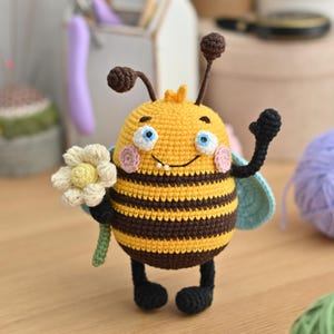 以下が含まれることがあります： 黄色と茶色の縞模様、青い羽根、白い花を持つ、かぎ針編みで作られた蜂の玩具。蜂は青い目とピンクの頬をした笑顔をしています。