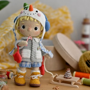Könnte beinhalten: Eine gehäkelte Puppe mit einem Schneemann-Hut und einer grauen Jacke, die einen Zuckerstock hält. Die Puppe steht auf einer Holzoberfläche mit einem kleinen Holzschlitten und Garn.