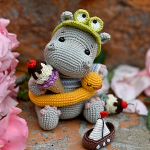 Op de afbeelding: Een gehaakte grijze nijlpaard met een groene kikkerhoed, met een ijsje met een kers erop. Een gele sjaal is om zijn nek gewikkeld en een kleine boot met een wit zeil staat in de buurt. Het nijlpaard is omringd door roze bloemen.