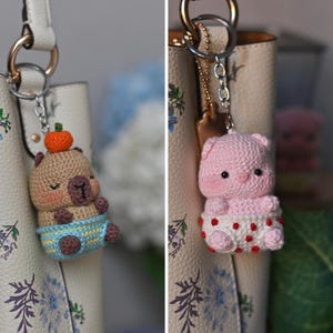 Puede incluir: Dos llaveros de ganchillo: uno con un oso marrón con sombrero de calabaza naranja y pañal azul, el otro un cerdo rosa con falda blanca de lunares rojos. Ambos están unidos a llaveros plateados.