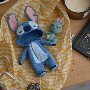 Puede incluir: Peluche de Stitch de crochet azul y blanco con orejas rosas y un mono azul con capucha. También se muestra un peluche de crochet verde más pequeño con un lazo rosa.