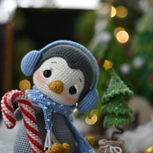 May include: A crocheted penguin toy wearing blue earmuffs and a blue and white scarf, holding a candy cane. The penguin is sitting next to a small crocheted Christmas tree.