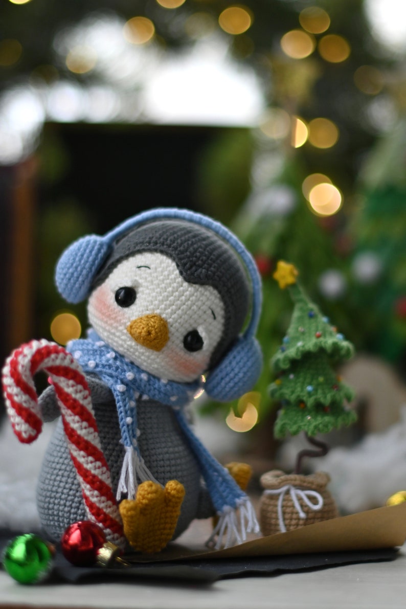 May include: A crocheted penguin toy wearing blue earmuffs and a blue and white scarf, holding a candy cane. The penguin is sitting next to a small crocheted Christmas tree.