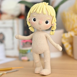 Puede incluir: Una muñeca de crochet con cabello rubio y un cuerpo beige. La muñeca está de pie sobre una superficie de madera.