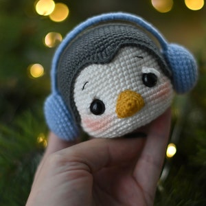 May include: A crocheted penguin toy wearing blue earmuffs. The penguin has black eyes, a yellow beak, and pink cheeks.