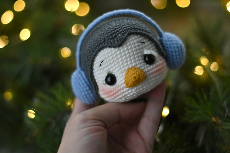 May include: A crocheted penguin toy wearing blue earmuffs. The penguin has black eyes, a yellow beak, and pink cheeks.