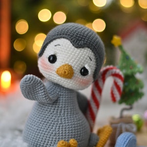 May include: A gray and white crocheted penguin toy holding a candy cane. The penguin has a yellow beak and black eyes. The toy is sitting on a brown surface with a small Christmas tree in the background.