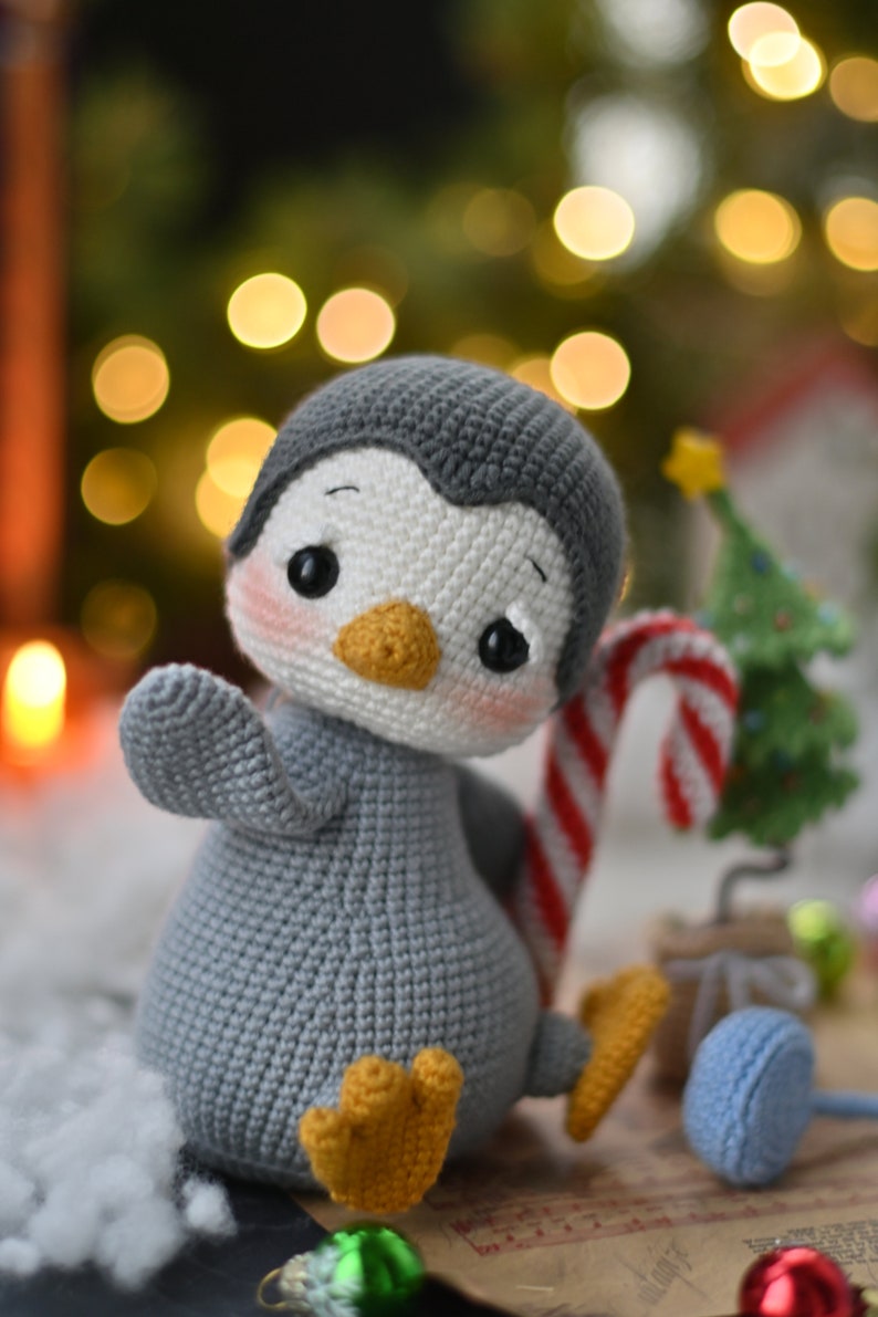 May include: A gray and white crocheted penguin toy holding a candy cane. The penguin has a yellow beak and black eyes. The toy is sitting on a brown surface with a small Christmas tree in the background.