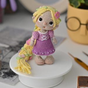 Può includere: Bambola all'uncinetto fatta a mano di una principessa con lunghi capelli biondi, che indossa un vestito viola con accenti rosa. La bambola è in piedi su una piattaforma bianca. La bambola è alta circa 15 cm.