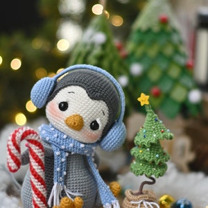 May include: A crocheted penguin wearing blue earmuffs and a blue and white scarf holds a candy cane. The penguin is sitting next to a small crocheted Christmas tree with a yellow star on top.