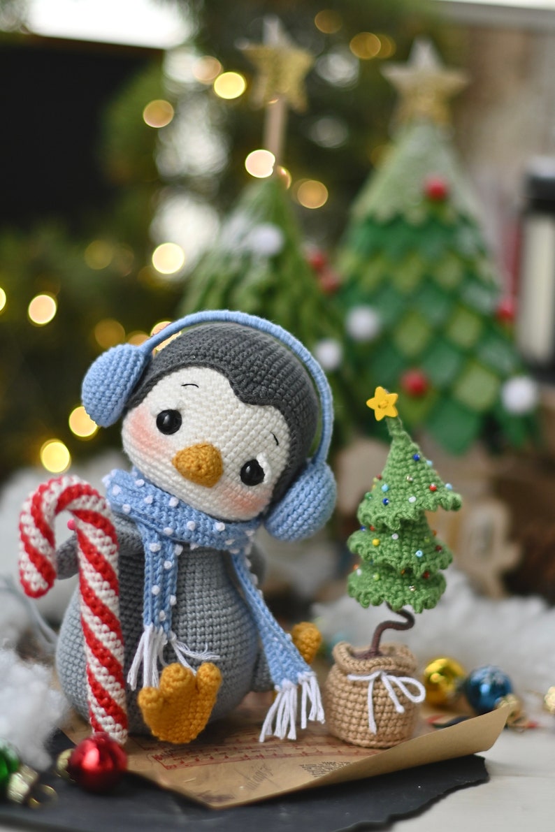 May include: A crocheted penguin wearing blue earmuffs and a blue and white scarf holds a candy cane. The penguin is sitting next to a small crocheted Christmas tree with a yellow star on top.