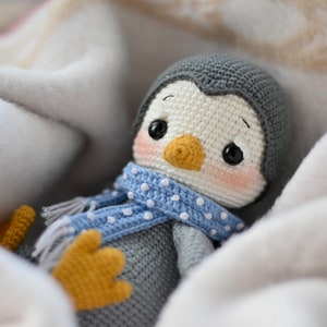 May include: A crocheted penguin toy wearing a blue and white polka dot scarf, nestled in a white blanket.