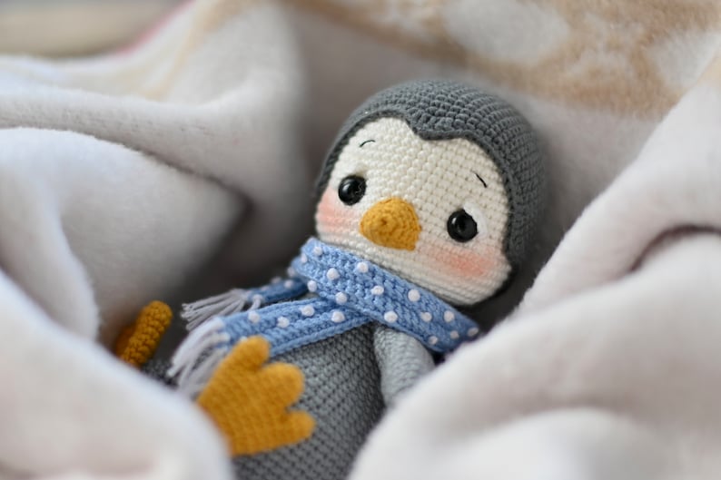 May include: A crocheted penguin toy wearing a blue and white polka dot scarf, nestled in a white blanket.