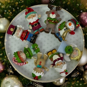 Puede incluir: Una colección de adornos navideños hechos a mano con ganchillo, que incluyen a Papá Noel, un reno, un muñeco de nieve y un elfo, dispuestos en un plato decorativo. Los adornos son de varios colores, como rojo, verde y blanco, y están rodeados de ramas de árbol de Navidad y bolas.