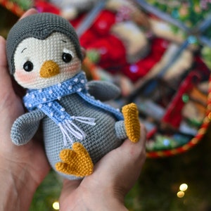 May include: A gray crocheted penguin wearing a blue and white polka dot scarf. The penguin has a white face with black eyes and a yellow beak.