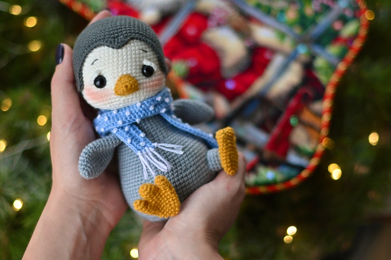 May include: A gray crocheted penguin wearing a blue and white polka dot scarf. The penguin has a white face with black eyes and a yellow beak.