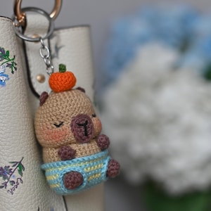 Puede incluir: Un llavero de ganchillo hecho a mano con un pequeño oso con una calabaza en la cabeza. El oso es de color marrón claro con mejillas rosadas, y lleva unos pantalones cortos a rayas azules y amarillas. Cuelga de una cadena plateada sujeta a un bolso blanco.