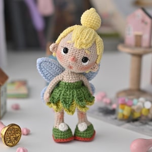 Puede incluir: Una muñeca de hada de ganchillo hecha a mano con un moño amarillo, cabello rubio y alas azules. La muñeca lleva una falda verde y zapatos verdes con detalles blancos. El hada tiene mejillas rosadas y ojos negros.