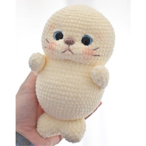 May include: A handmade, cream-colored crochet seal plushie. The seal has blue eyes, a brown nose, and blush-pink cheeks. It has small flipper-like arms and a tail. The plushie is made with a textured yarn, giving it a soft appearance.