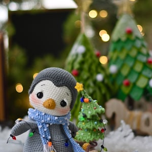 May include: A gray crocheted penguin wearing a blue and white polka dot scarf and holding a string of Christmas lights. The penguin is standing next to a small green crocheted Christmas tree with lights and a brown crocheted gift bag.