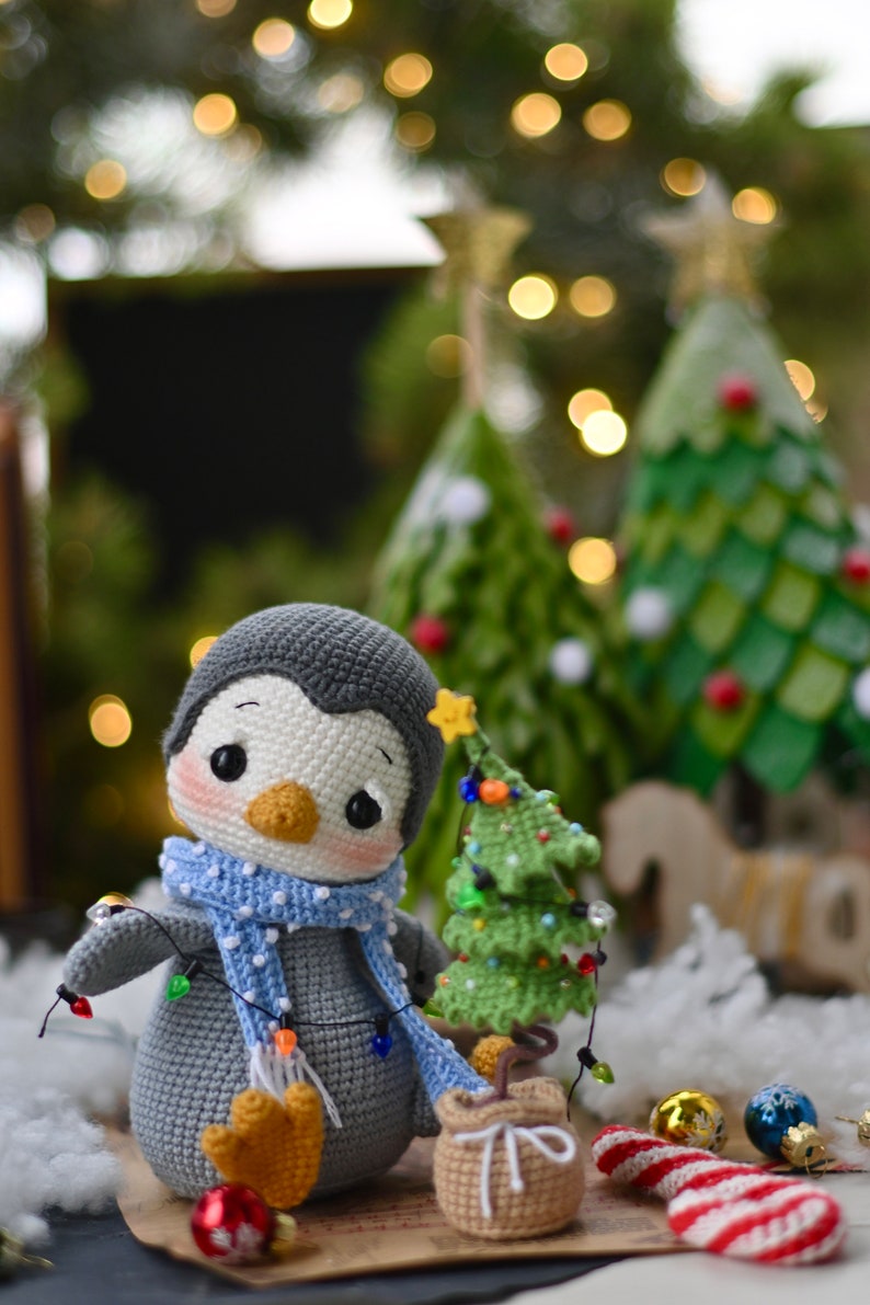 May include: A gray crocheted penguin wearing a blue and white polka dot scarf and holding a string of Christmas lights. The penguin is standing next to a small green crocheted Christmas tree with lights and a brown crocheted gift bag.
