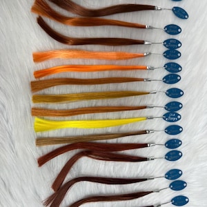 May include: A collection of hair swatches in various shades of brown, orange, and yellow. Each swatch is attached to a small metal loop with a blue tag. The tags have white text. The swatches are arranged on a white, textured surface.